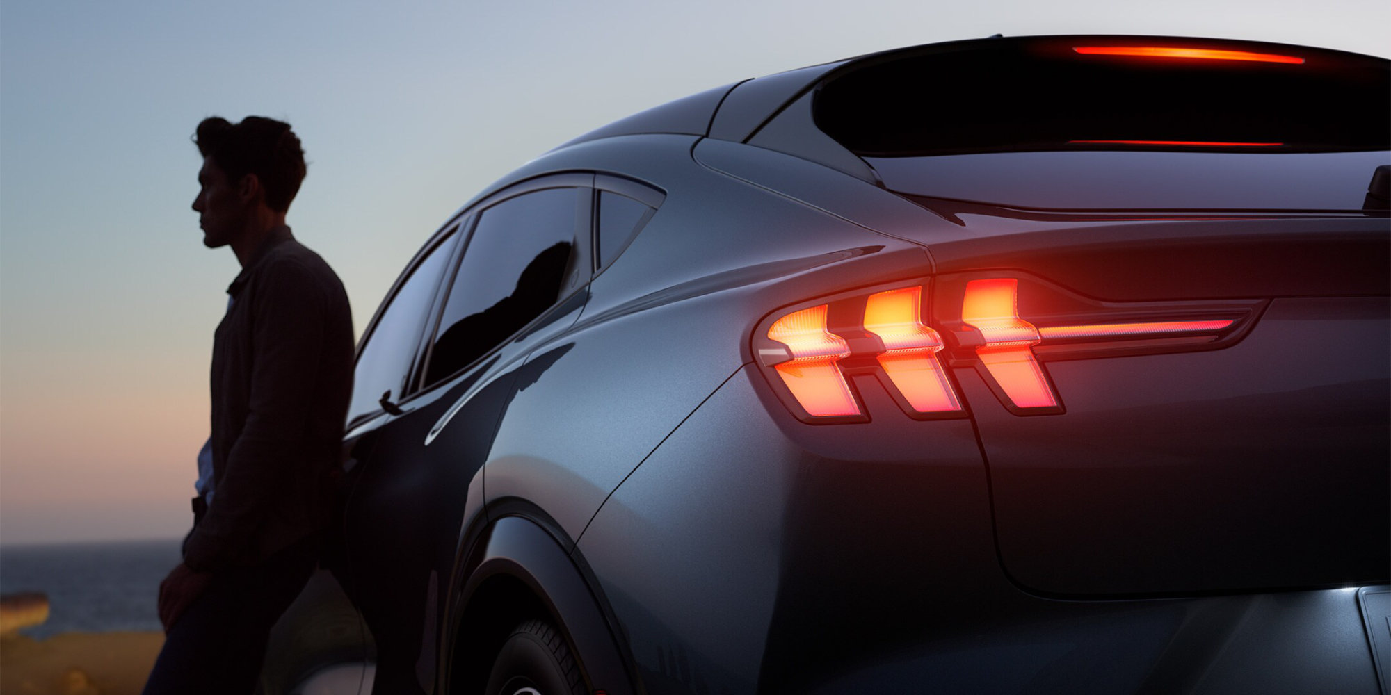 Ford Mustang Mach-E in Grau. Mann lehnt in Abenddämmerung am Fahrzeug